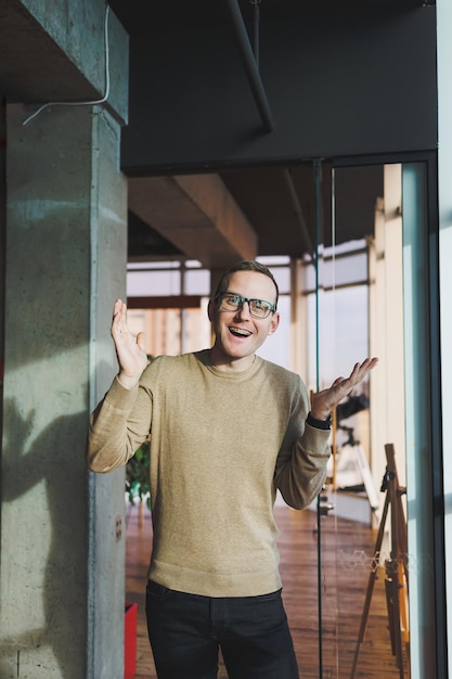 A young man in glasses and casual clothes stands in a modern workspace Male freelancer works remotely