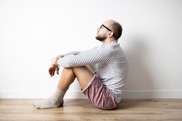 A young man in glasses crosses his legs sitting on a wooden floor looking up