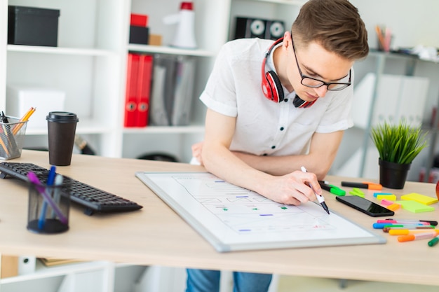 A young man in glasses stands near a computer desk. 