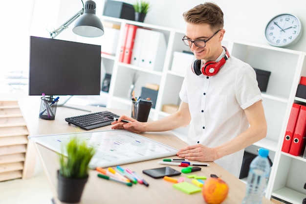 A young man in glasses stands near a computer desk.