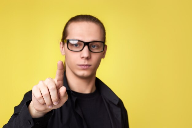 Young man in glasses touching virtual screen on yellow background. focus on finger