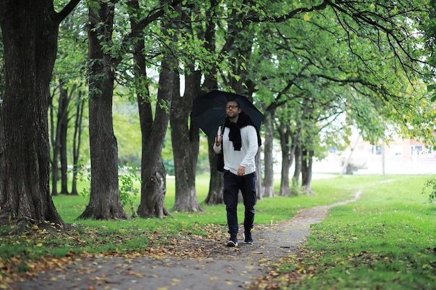 A young man in glasses walks in the park with an umbrella during the rain.