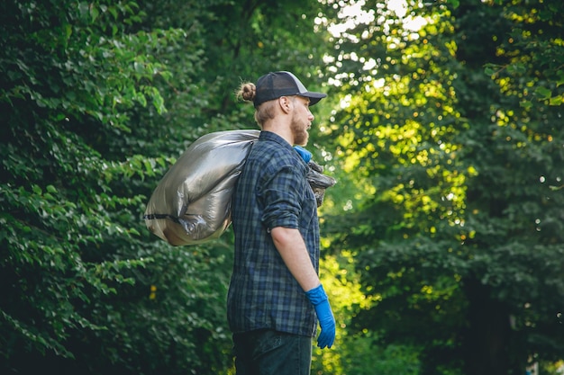 A young man in gloves and with a garbage bag cleans up in the forest