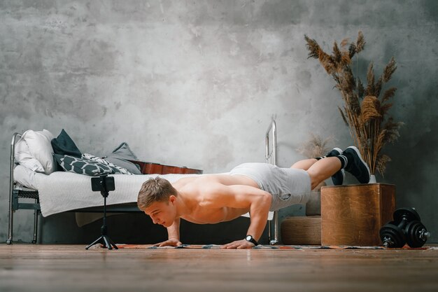 The young man goes in for sports at home. The athlete does push-ups and watches a movie and studies from a phone  in the bedroom, in the background a bed, a vase, a carpet