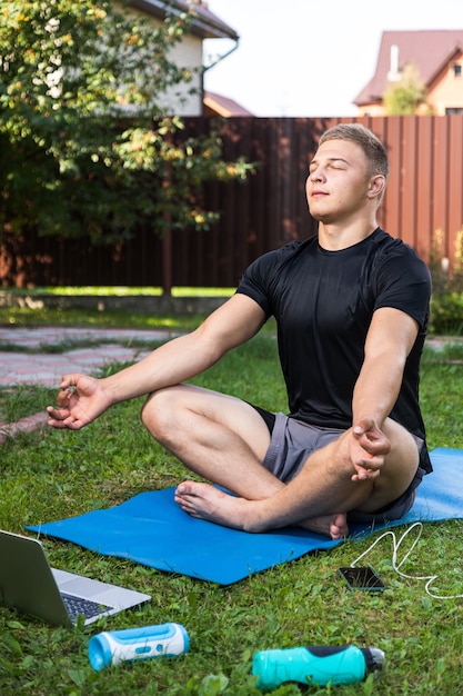 The young man goes in for sports at home in  backyard in summer day. Cheerful sportsman with blonde hair meditates in lotus position, resting on the mat  in garden