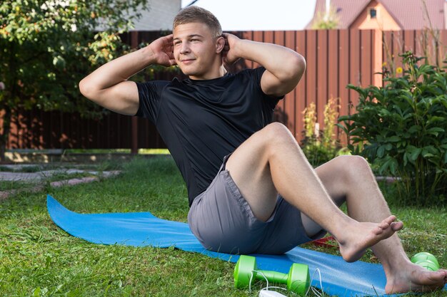 Young man goes in for sports  at home in  backyard in summer day, training online. The athlete makes the press on the mat  in garden