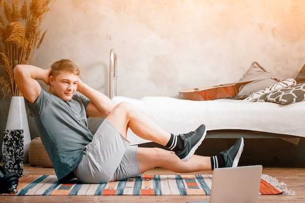 Photo the young man goes in for sports at home. cheerful sportsman with blond hair makes the press in the bedroom, next to there is a laptop with online training