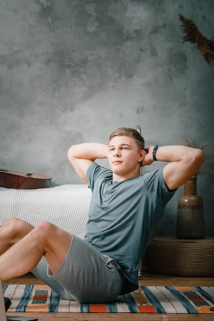 Young man goes in for sports at home, training online. The athlete makes the press, smiles, there is an open laptop, bed, vase, carpet nearby.