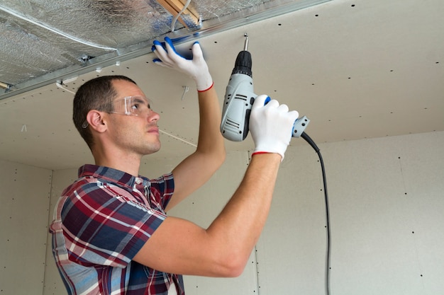 Young man in goggles fixing drywall suspended ceiling to metal frame using electrical screwdriver on ceiling insulated with shiny aluminum foil. Renovation, construction, do it yourself concept.