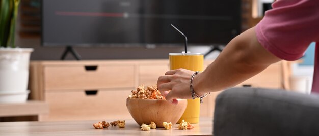 Young man grabbing popcorn from a wooden bowl and watching TV at home