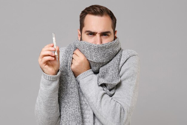 Young man in gray sweater isolated on grey background studio portrait. Healthy lifestyle ill sick disease treatment cold season concept. Mock up copy space. Hold thermometer covering mouth with scarf.