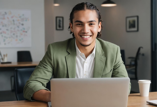 Молодой человек в зеленой куртке работает на своем ноутбуке улыбается в сторону камеры офисной среды