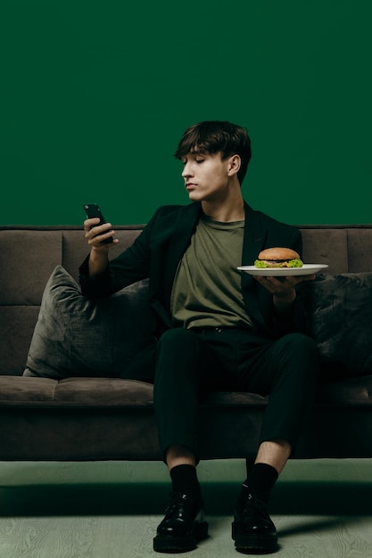 A young man in a green stylish suit is eating a burger while
sitting on a sofa on a green background fast food concept