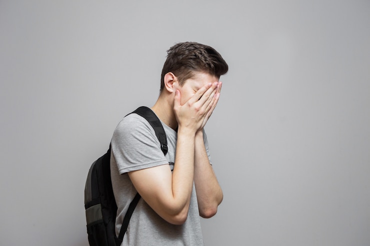 灰色のシャツを着た若い男は、彼の手で顔を覆っています。灰色の背景の男