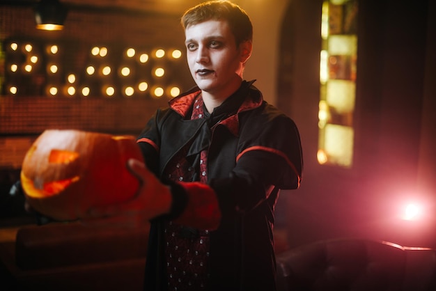 A young man in a Halloween costume of Count Dracula holds a carved pumpkin on his outstretched arm