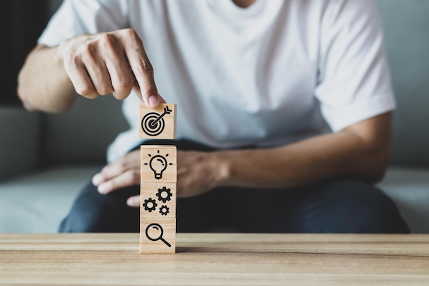 Young man hand putting wooden Blocks with icon of goal gearwheels lightbulb and magnifying glass Demonstrates having goals developing looking for new ideas to develop the business to grow