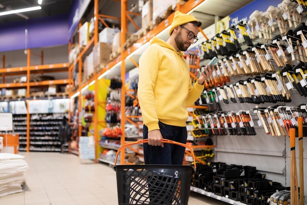 Photo a young man in a hardware store next to a rack with hammers and axes looks at the smartphone screen