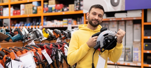 A young man in a hardware store in the tool department embracing a circular saw for wood