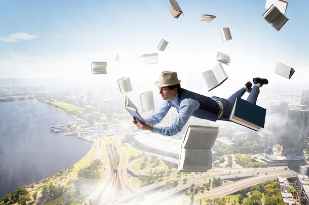 Young man in hat and casual clothes levitating over city with books around. Mixed media