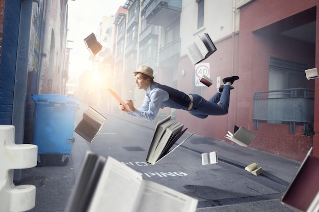 帽子とカジュアルな服を着た若い男が、本や周りに物を置いて街の上を浮揚している。ミクストメディア