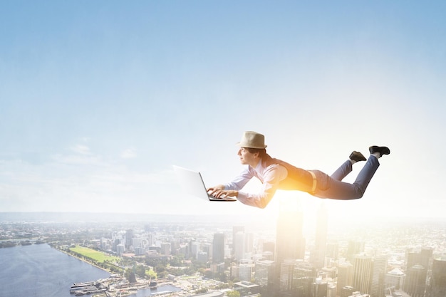 Young man in hat and casual clothes levitating over city with laptop. Mixed media