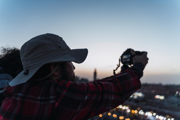 Photo young man in hat takes pictures of a city at dusk