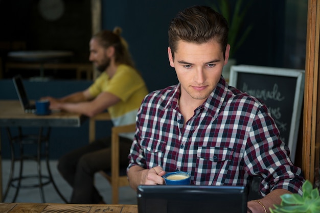 Young man having coffee while using digital tablet in cafeteria