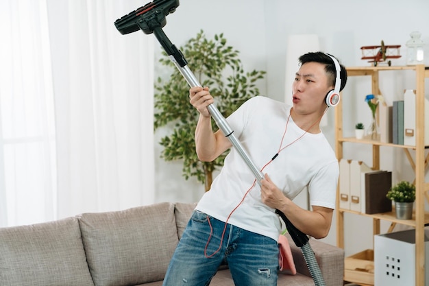 Фото Молодой человек весело убирает дом с пылесосом, танцующим как гитарист. азиатский корейский парень слушает музыку в наушниках и наслаждается пением на диване. мужское домашнее хозяйство с пылесосом развлекается