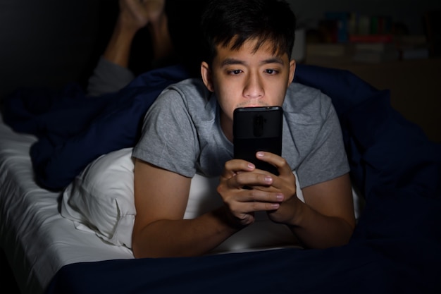 Young man having sore and tired eyes when using smartphone while lying in bed at night