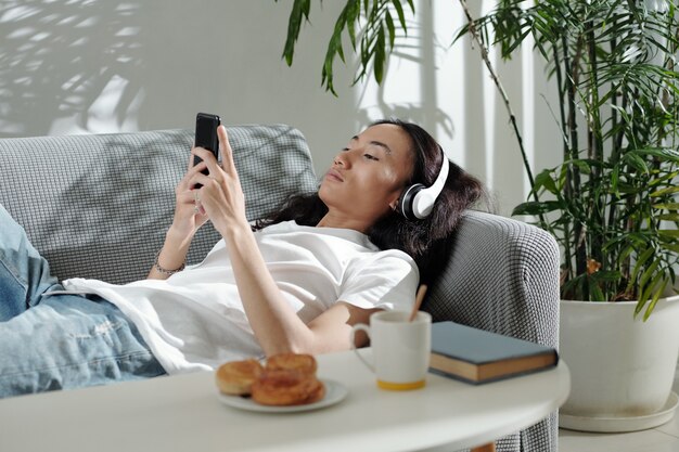 Young man in headphones lying on sofa listening to music and texting friend or playing addictive gam...