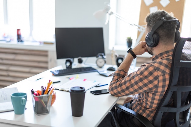 A young man in headphones sits at a table in the office and looks at the monitor. The picture is taken from behind the shoulder.