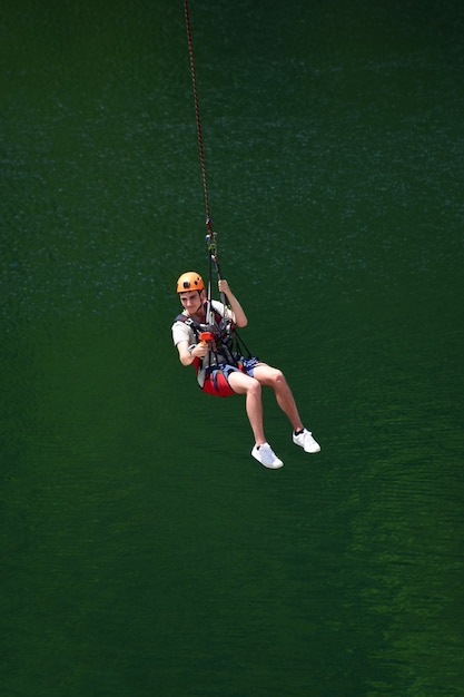 Un giovane con un casco è saltato dal bungee jumping e ora è appeso a una corda, oscillando e riprendendosi su una videocamera sportiva su uno sfondo sfocato d'acqua
