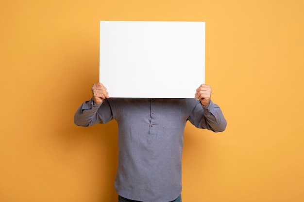 Young man hiding face behind blank white placard over yellow background