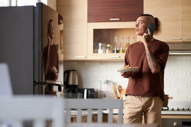 Young man hoding plate with sandwiches when talking on phone with friend or coworker