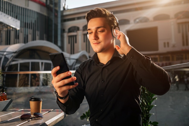 Young man hold phone in hand. he is outside at table. guy listen to music. it is sunny outside.