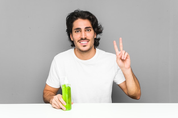 Young man holding an aloe vera bottle showing victory sign and smiling broadly.