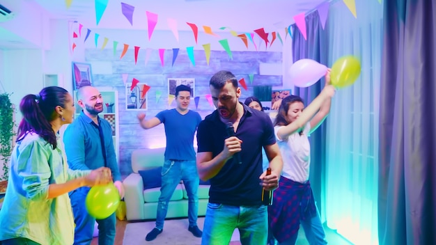 Young man holding a beer bottle at a party and singing karaoke. Group of people dancing at wild college party with neon lights.