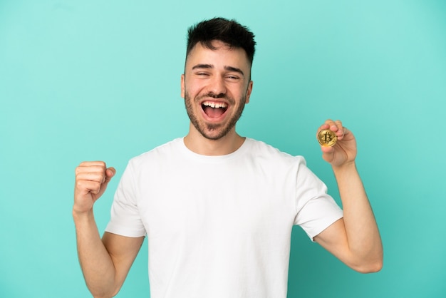 Young man holding a Bitcoin isolated on blue background celebrating a victory in winner position