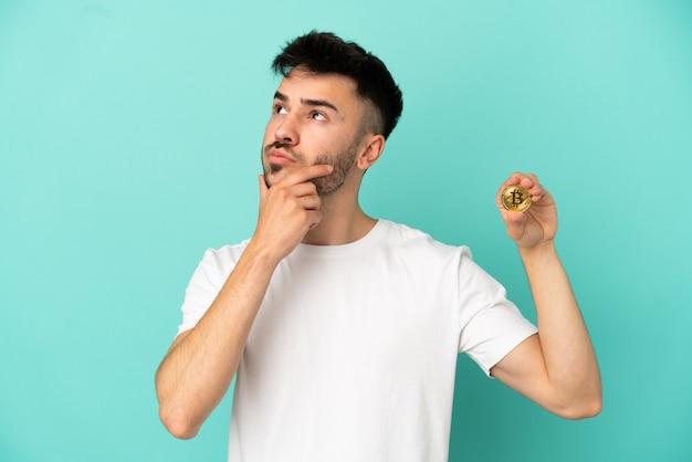 Young man holding a Bitcoin isolated on blue background having doubts