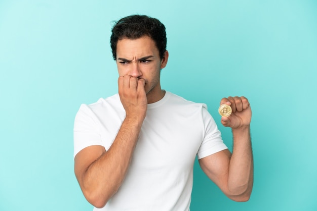 Young man holding a Bitcoin over isolated blue background having doubts