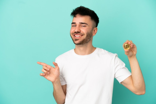 Young man holding a Bitcoin isolated on blue background pointing finger to the side