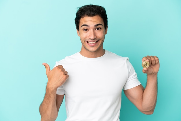 Young man holding a Bitcoin over isolated blue background pointing to the side to present a product