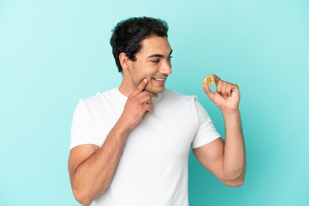 Young man holding a Bitcoin over isolated blue background thinking an idea while looking up