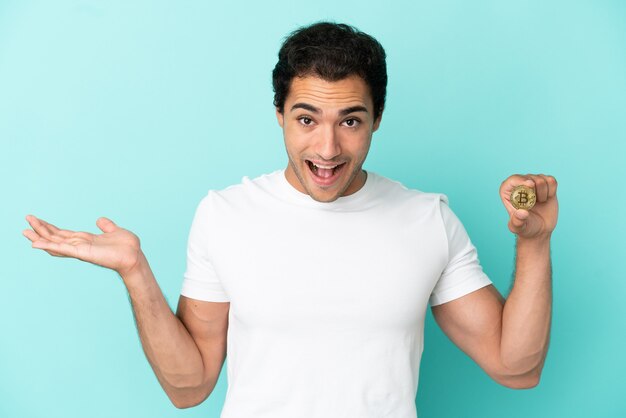 Young man holding a Bitcoin over isolated blue background with shocked facial expression