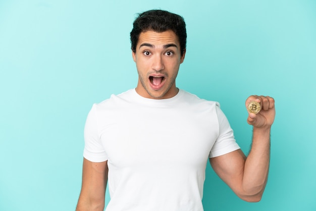 Young man holding a Bitcoin over isolated blue background with surprise facial expression