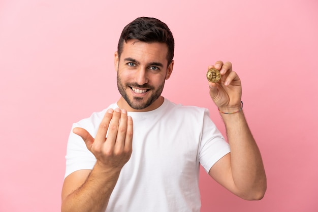 Young man holding a Bitcoin isolated on pink background inviting to come with hand. Happy that you came