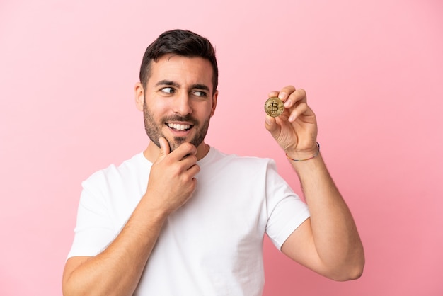 Young man holding a Bitcoin isolated on pink background looking to the side and smiling