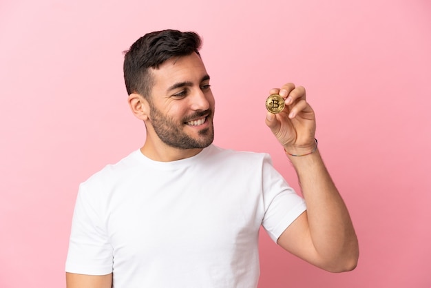 Young man holding a Bitcoin isolated on pink background looking to the side and smiling