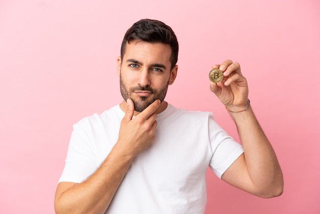Young man holding a Bitcoin isolated on pink background thinking