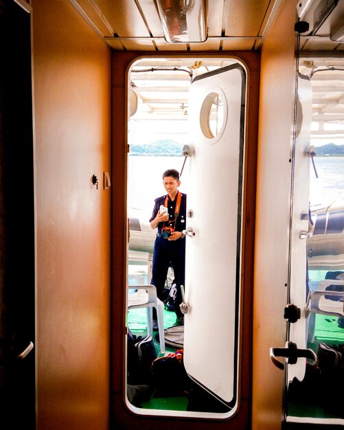 Young man holding camera and smart phone in boat seen through doorway